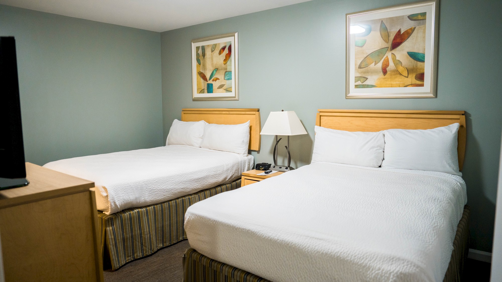 The image shows a hotel room with two double beds, white bedding, a lamp, and abstract art on the walls.