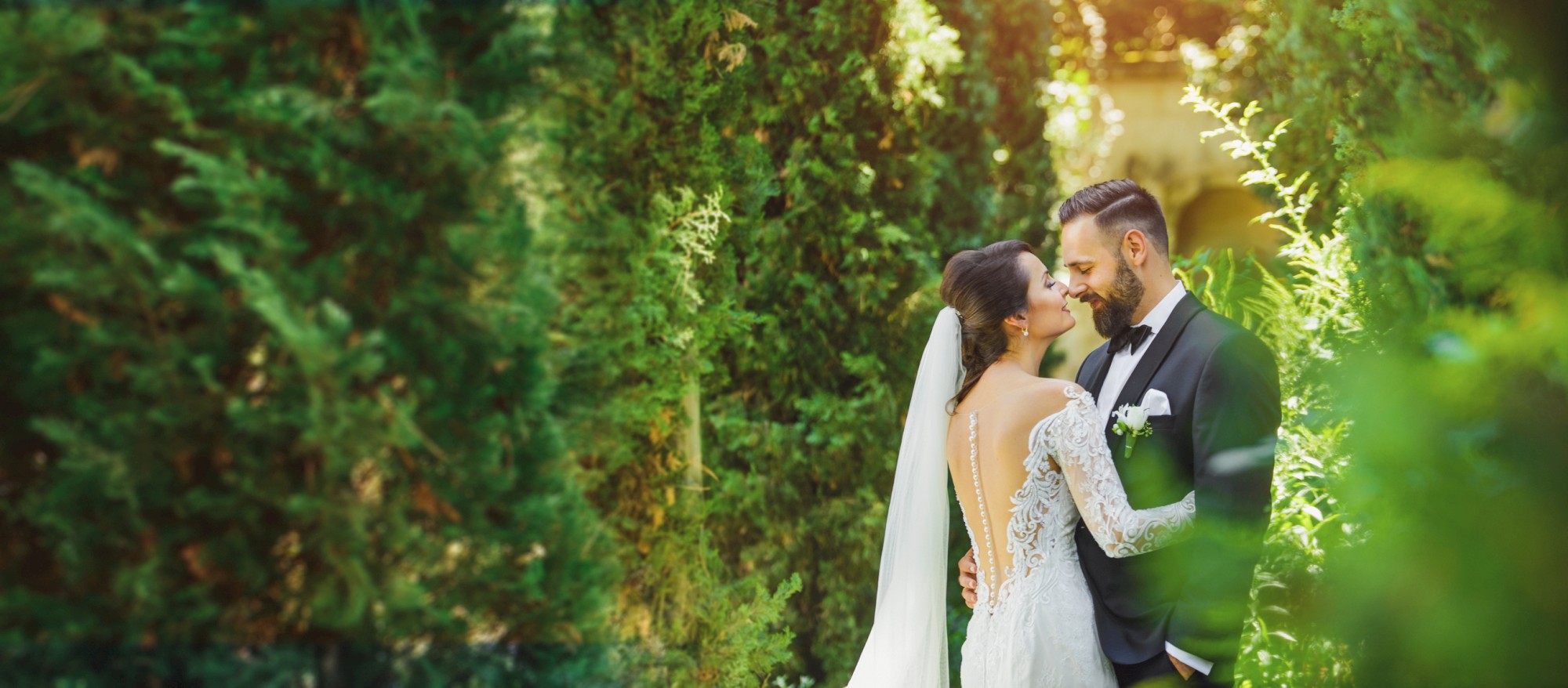 A bride and groom stand closely together in a lush garden setting, surrounded by greenery and sunlight peeking through the trees.