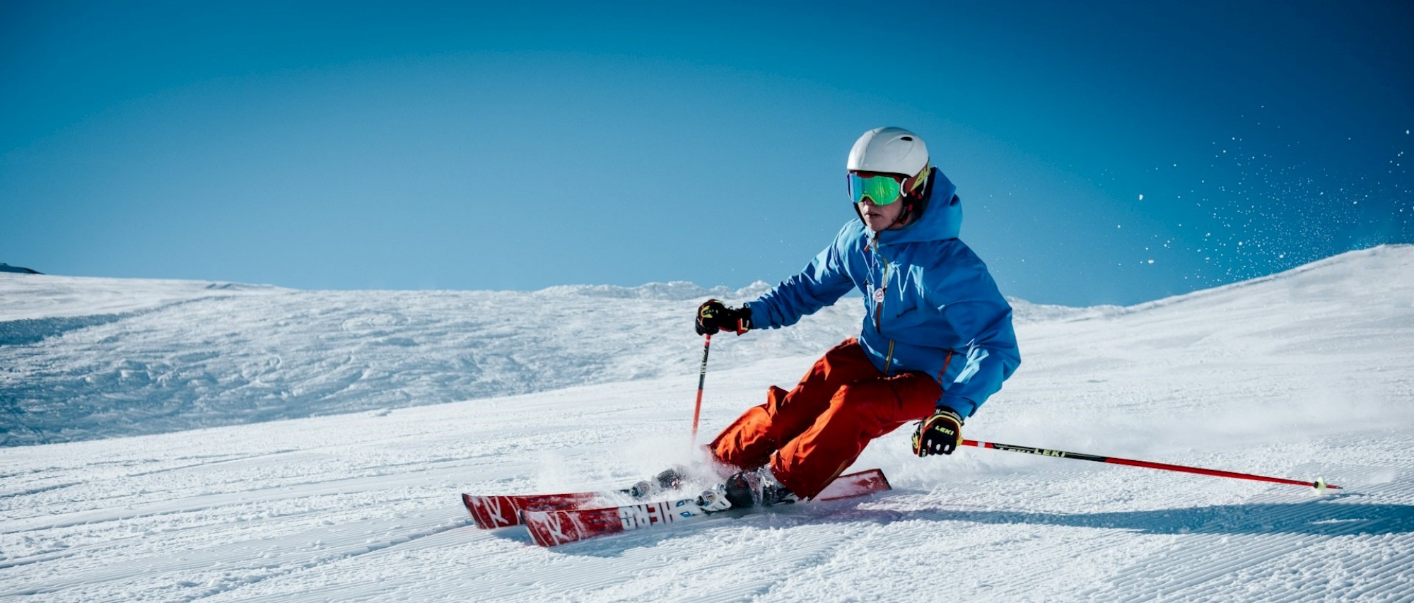 A person is skiing downhill on a snowy slope, wearing a blue jacket, red pants, and a helmet with goggles, under a clear blue sky.