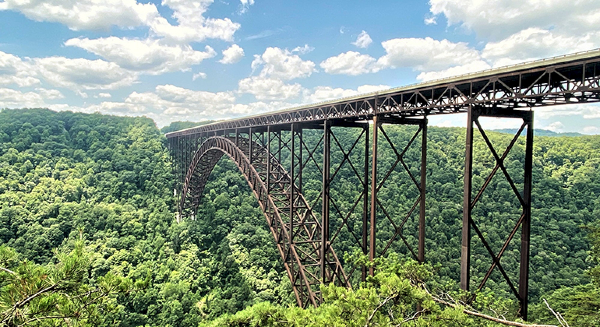 The image shows a large arched steel bridge spanning a lush, forested gorge under a partly cloudy blue sky.