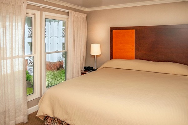 A cozy bedroom with a large bed, wooden headboard, bedside lamp, and sheer curtains covering the window.