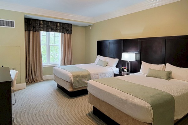 The image shows a hotel room with two double beds, a window with curtains, a TV on a dresser, and a lamp on a bedside table.