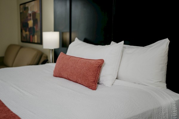 A neatly made bed with white pillows, a red throw pillow, and a red blanket in a dimly lit room with a lamp and wall art.