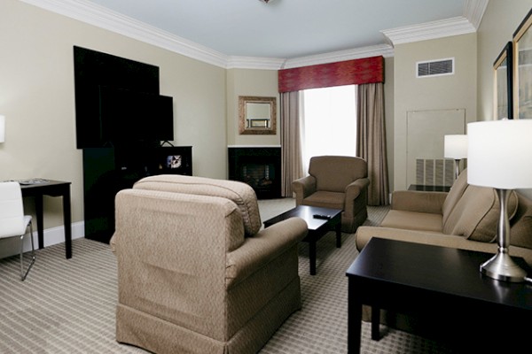 A living room with beige armchairs, a desk with a chair, lamps, and a TV. The room has a window with curtains and carpet flooring.