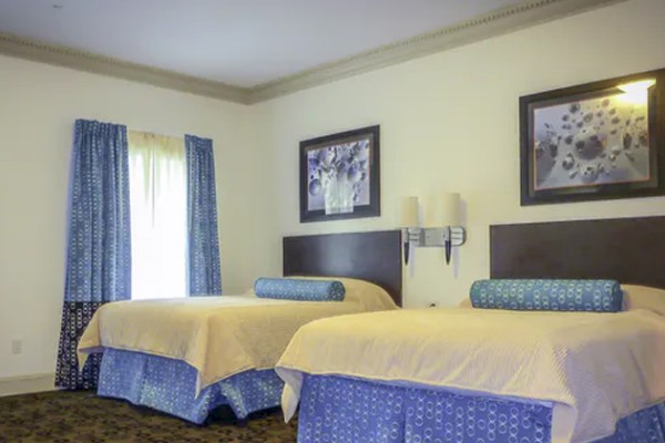 The image shows a hotel room with two double beds, blue bedding, curtains, framed pictures, and a wall lamp above a nightstand.