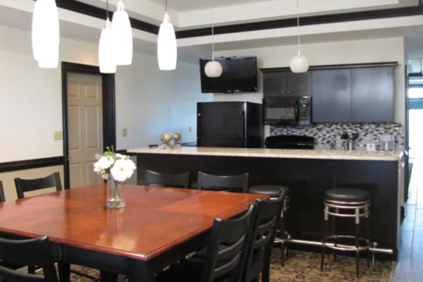 The image shows a dining area with a wooden table, black chairs, hanging lights, and a kitchen with bar stools and a TV on the wall.