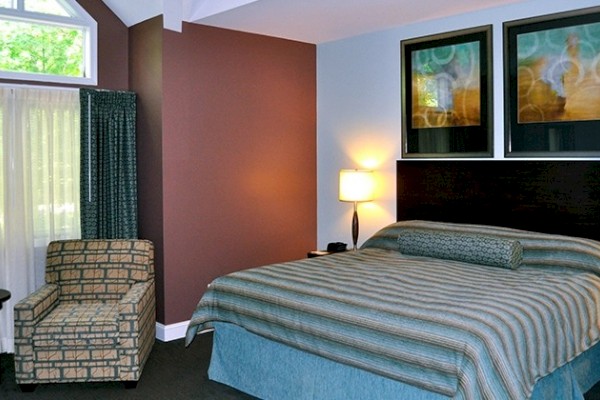 A cozy bedroom with a large bed, striped bedding, an armchair, two framed artworks, and a window with curtains.