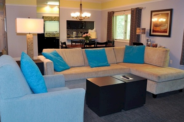 A cozy living room with a beige sectional sofa, blue pillows, a lamp, coffee table, and open view into the dining area and kitchen.