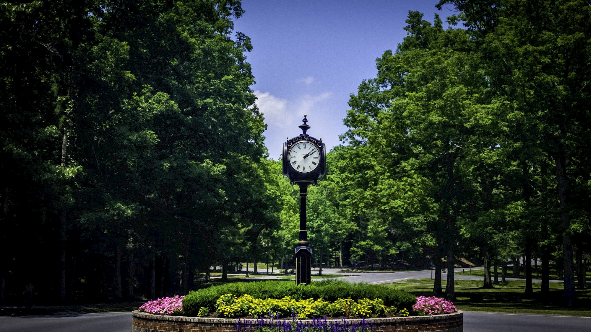 An ornate clock stands at the center of a landscaped roundabout, surrounded by vibrant flowers and lush green trees under a clear blue sky.