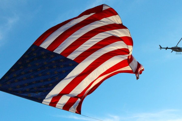 A large American flag is flying in the sky, with a helicopter nearby in the background.