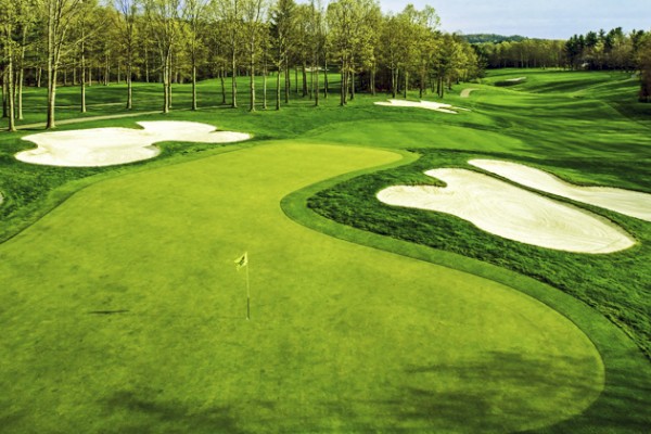 The image shows a golf course with a green, sand bunkers, and surrounding trees. The sky is clear, and the landscape appears lush and well-maintained.