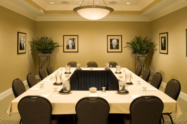 The image shows a conference room with a U-shaped table setup, framed portraits on the walls, and plants on the sides, under ceiling lights.