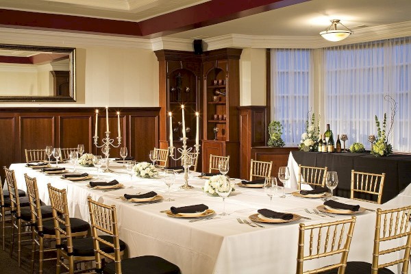 Elegantly set dining table with white tablecloth, gold chairs, black napkins, candelabras, and floral arrangements in a wood-paneled room.