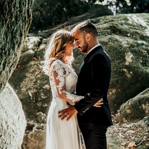 A couple in wedding attire embraces in a rocky, outdoor setting, surrounded by lush greenery.