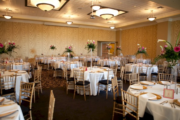 A banquet hall set with round tables, white tablecloths, golden chairs, and floral centerpieces, ready for an event or celebration.