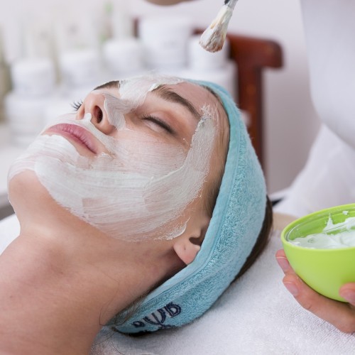 A person is receiving a facial treatment with a mask being applied by a professional using a brush, while holding a green bowl with cream.