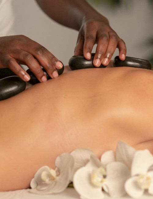 A person is receiving a hot stone massage, with black stones placed on their back alongside white flowers for a relaxing experience.