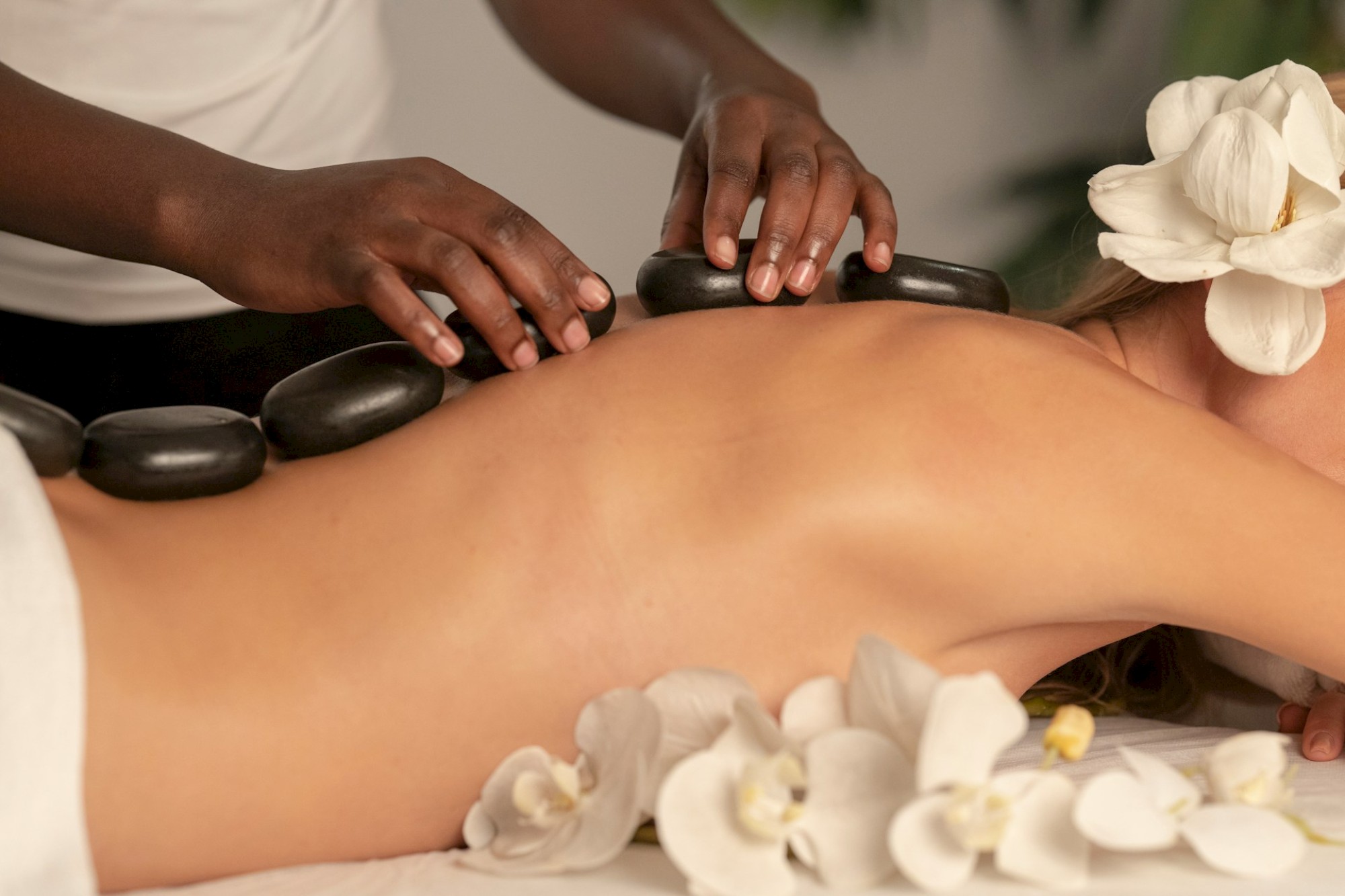 A person is receiving a hot stone massage on their back, surrounded by white flowers, creating a relaxing spa atmosphere.