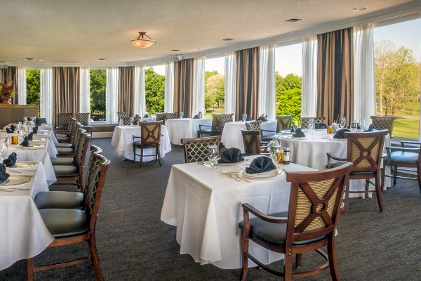 The image shows an elegant dining room with neatly set tables, large windows, and a view of greenery outside, creating a bright atmosphere.