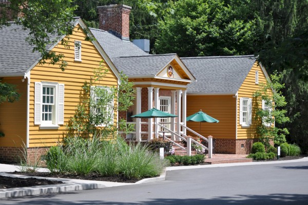 A quaint, yellow wooden building with green umbrellas on a porch, surrounded by lush greenery and trees, on a clear day.