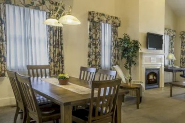 A cozy dining area with a wooden table, chairs, floral curtains, a fireplace with a TV above, and a small desk area in the corner.