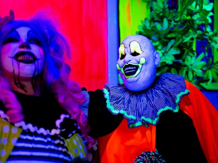 Two colorful clowns with face paint in a vibrant setting, surrounded by neon lights and greenery, creating a lively atmosphere.