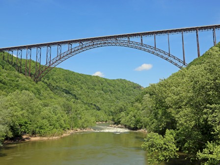 A steel arch bridge spans a lush, green valley with a river flowing below under a clear blue sky.