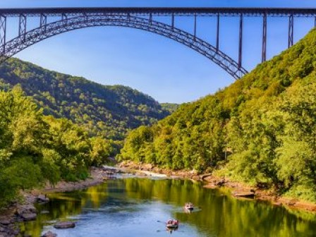 A large arch bridge spans over a lush green river valley with people kayaking on the water below, surrounded by dense forested hills.