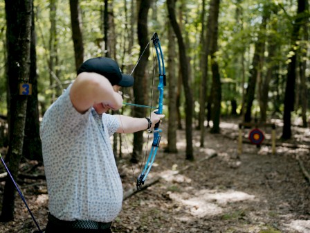 A person is aiming a bow and arrow in a wooded area with a target in the background.