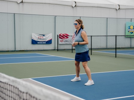 A woman is standing on a tennis court wearing sports attire and sunglasses.