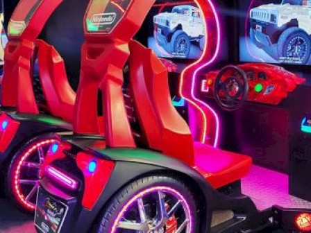 This image shows a colorful arcade racing game setup with two red seats, steering wheels, and screens displaying racing scenes.