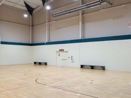 The image shows an indoor basketball court with wooden flooring, high ceilings, and basketball hoops on each end.