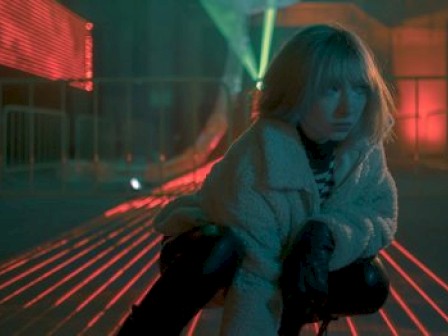 A person in a coat is crouching in a dimly lit, futuristic setting with neon lights and metal railings.