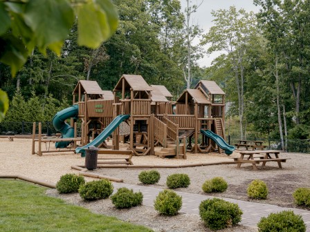 The image shows a playground with wooden structures, slides, and a sitting area, surrounded by trees and landscaping.