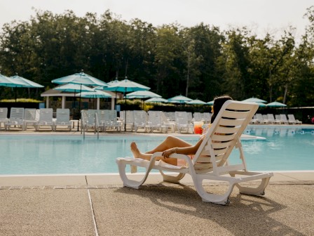 A person relaxes on a lounge chair beside a swimming pool with umbrellas and trees in the background.