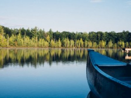 A canoe on a calm lake with a forested shoreline and clear blue sky in the background, reflecting serene and peaceful scenery.