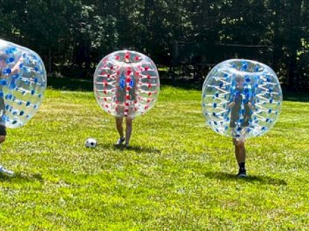 Three people playing bubble soccer on a grassy field, each inside a transparent inflatable ball.