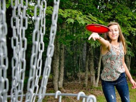 A woman is throwing a red disc towards a disc golf basket in a wooded area.