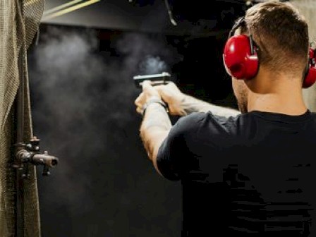 A person is wearing ear protection, aiming and shooting a handgun at an indoor firing range, with smoke visible around the gun.