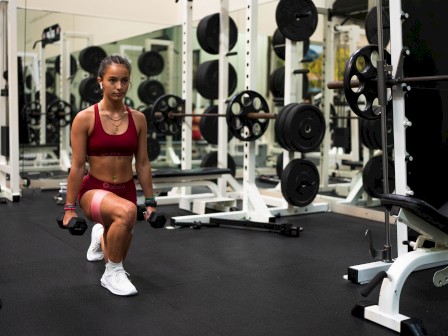 A person in workout attire is doing lunges with dumbbells in a gym filled with equipment and weights.