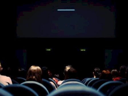 People sitting in a dimly lit movie theater, facing the screen, with rows of seats visible.