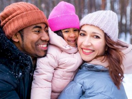 A happy family in winter clothing stands together, smiling in a snowy outdoor setting.