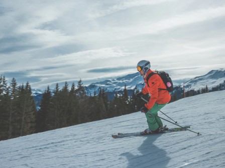 A person skiing downhill on a snowy slope with mountains and trees in the background, wearing bright gear and a helmet.