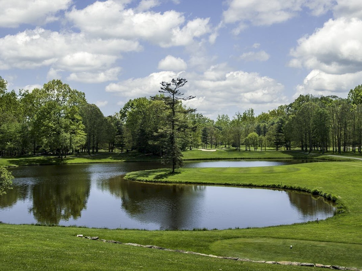 A lush golf course featuring a serene pond surrounded by trees and a neatly trimmed green landscape under a partly cloudy sky.