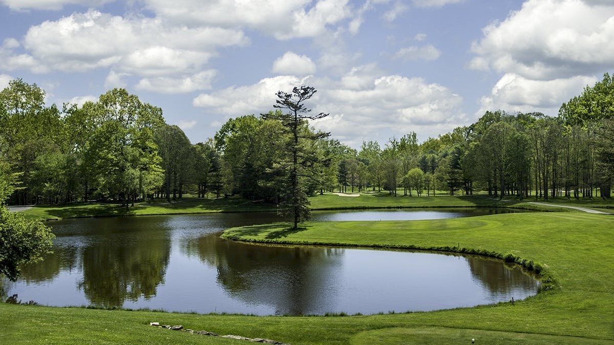 A lush golf course featuring a serene pond surrounded by trees and a neatly trimmed green landscape under a partly cloudy sky.