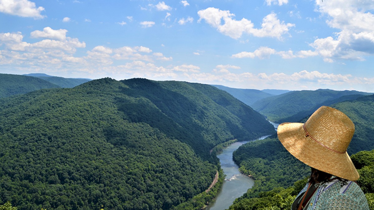 A person in a large straw hat overlooks a river winding through lush, green mountains under a partly cloudy blue sky.