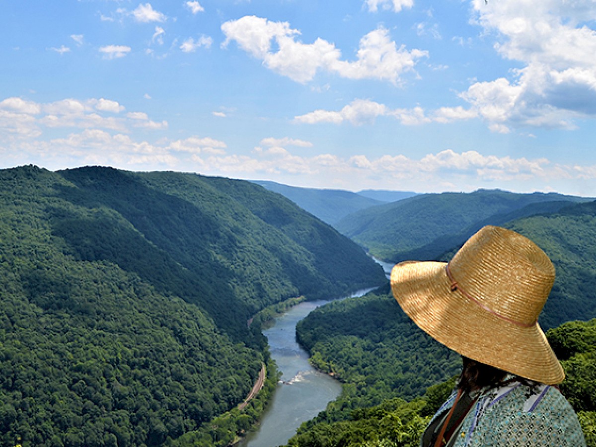 A person in a large straw hat overlooks a river winding through lush, green mountains under a partly cloudy blue sky.