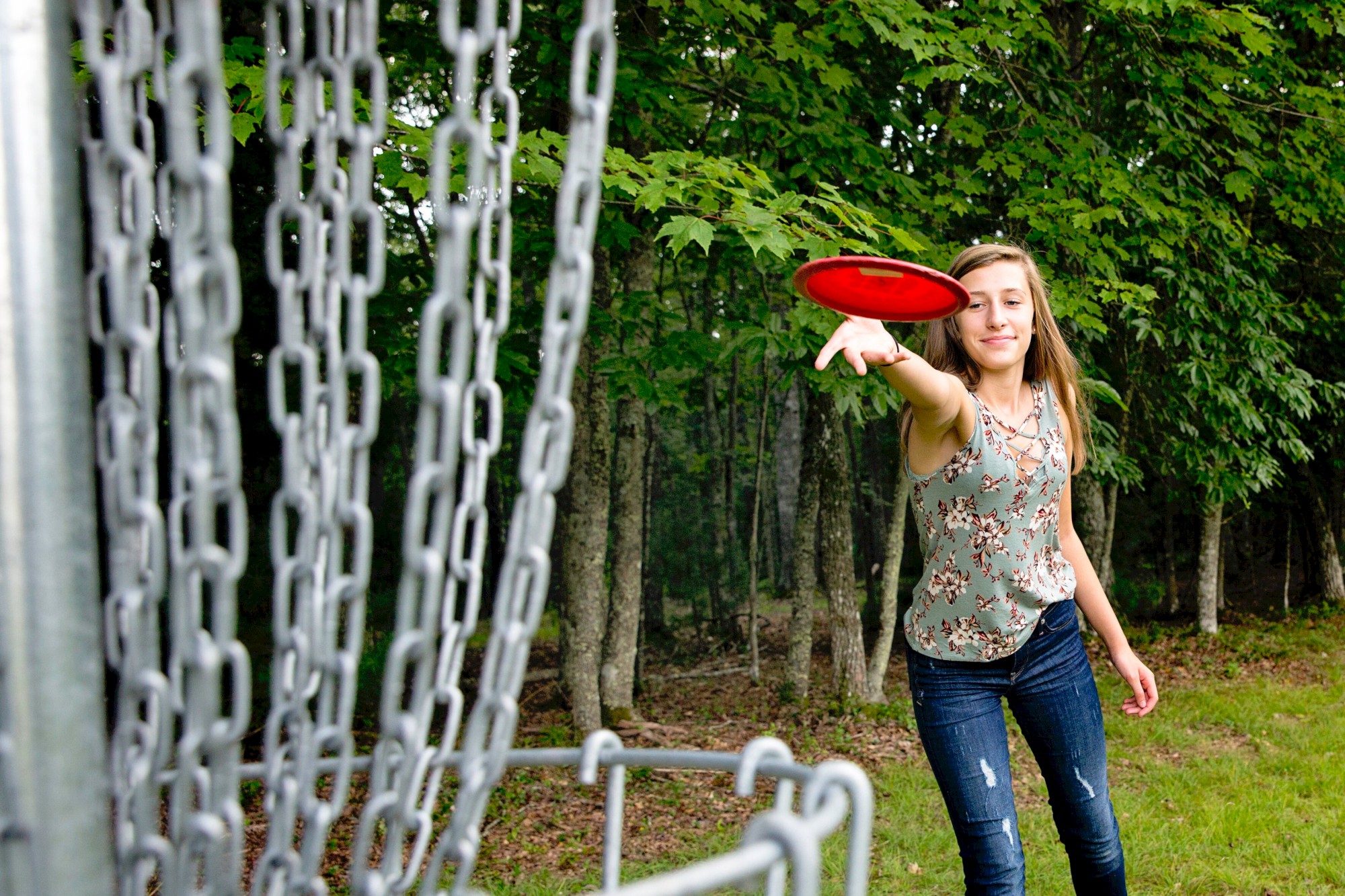 A person is playing disc golf, aiming a red frisbee towards a metal basket in a wooded area.