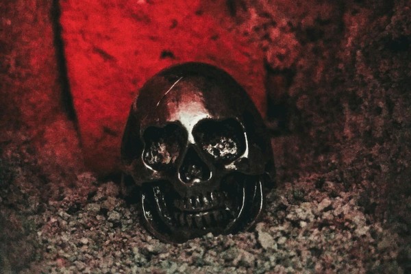 A metallic skull placed on gravel is illuminated by a red light in a dark, rocky setting.