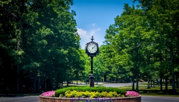 A roundabout with a vintage clock in the center, surrounded by a colorful flower bed and lush green trees under a clear blue sky.